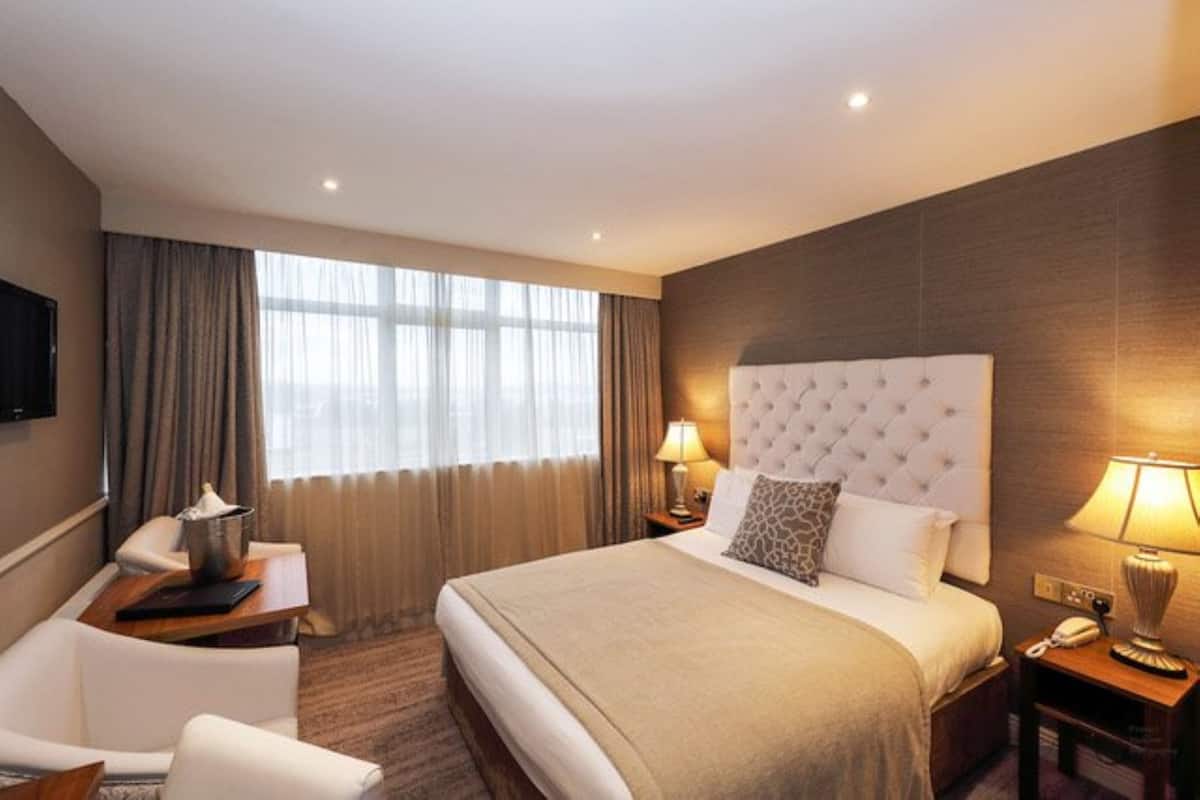 A hotel room with a large bed, a desk with a chair, a TV mounted on the wall, and a window with curtains.