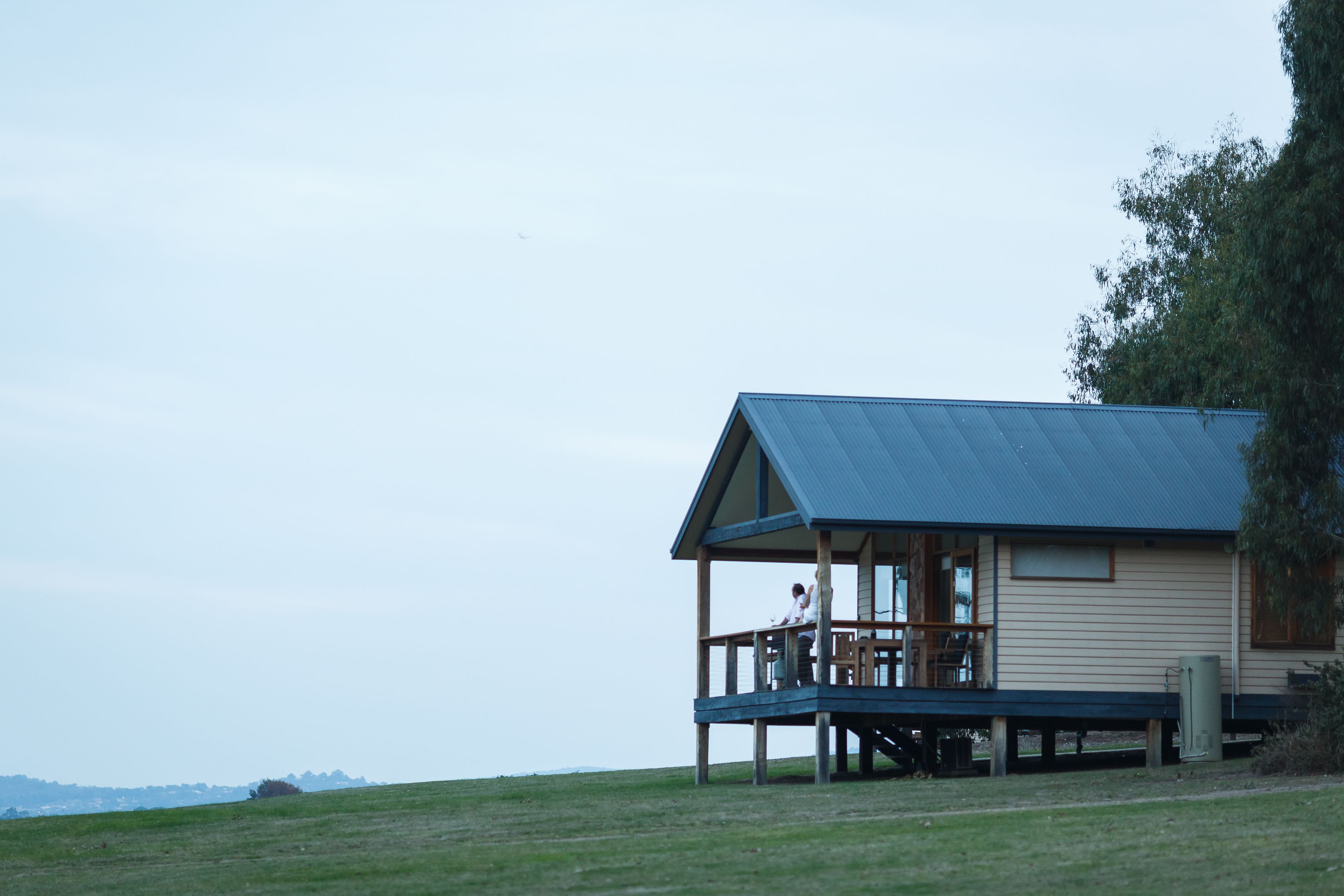 A wooden cabin with a balcony, situated on a grassy hill with a view of the landscape.