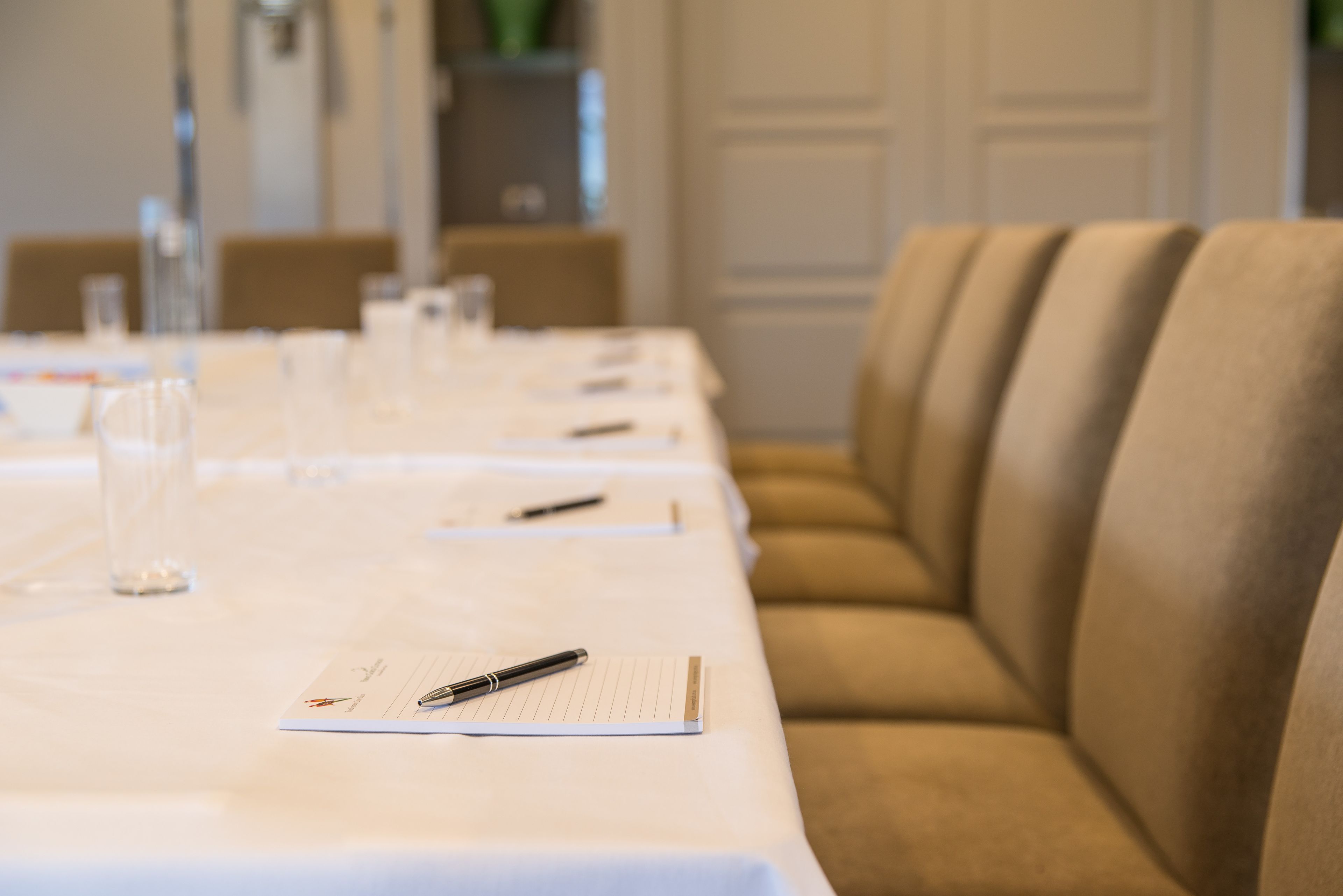 Une table de conférence avec des blocs-notes et des stylos, un verre d’eau, et un canapé beige en arrière-plan.