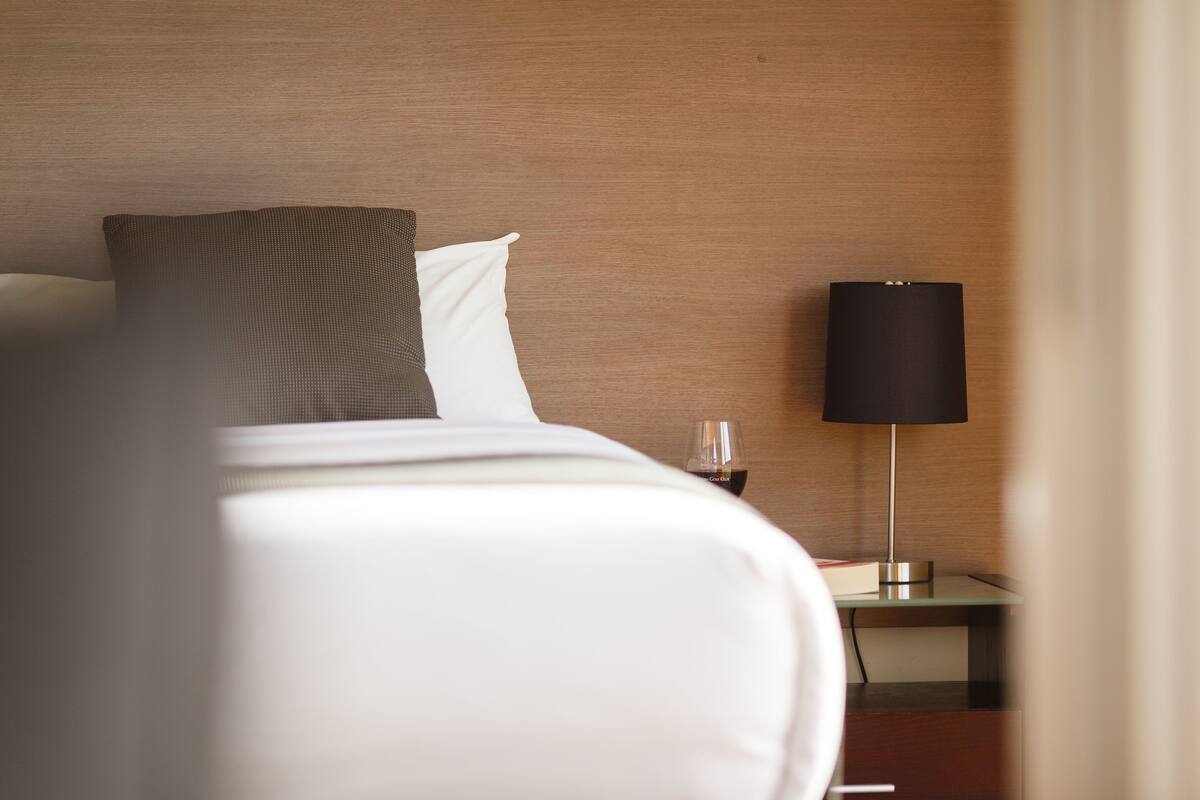A neatly made bed with white and brown bedding, a bedside table with a glass of red wine, and a modern lamp.