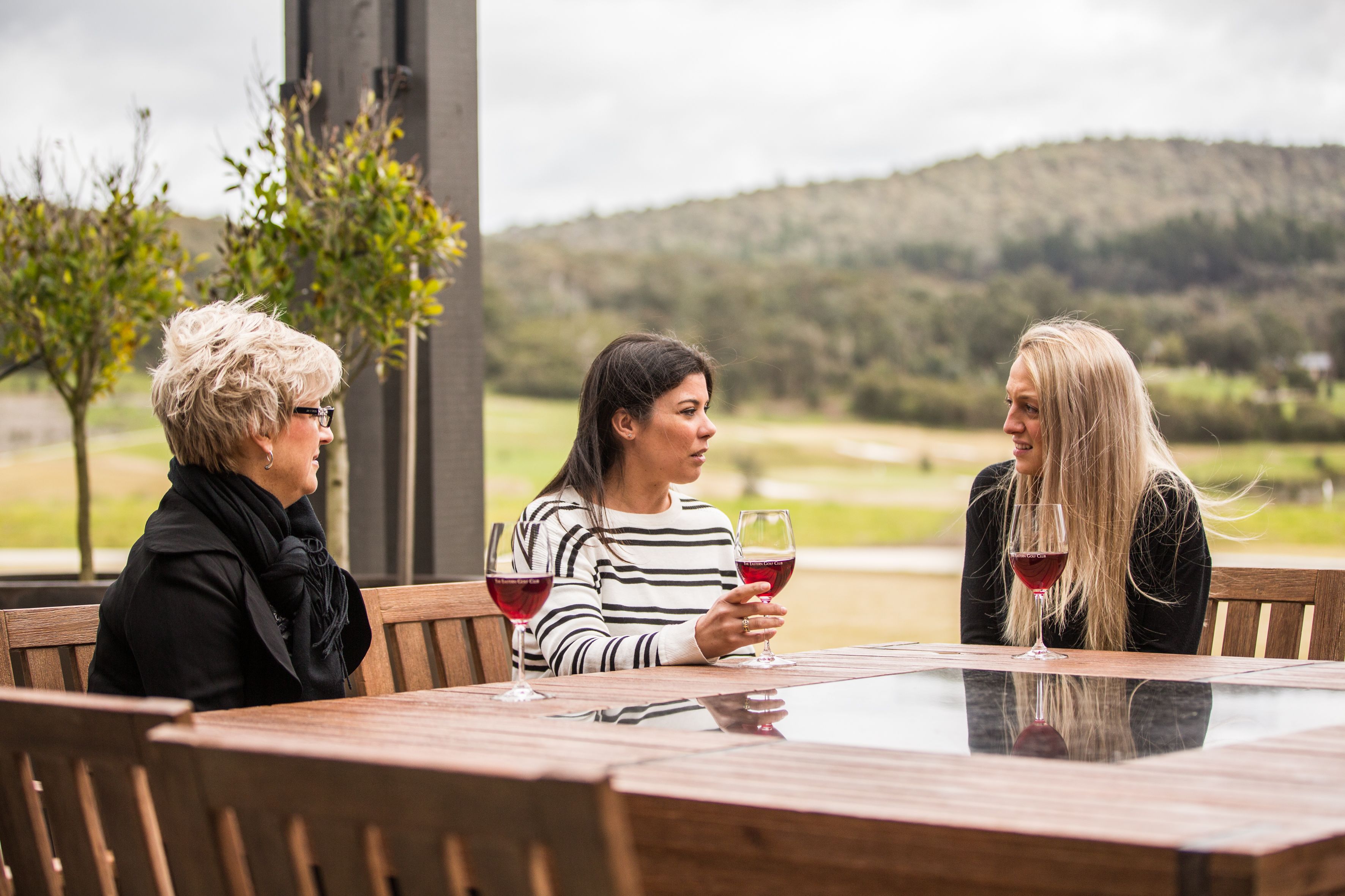 Three people seated at a wooden table with wine glasses, outdoors with a view of trees and hills.