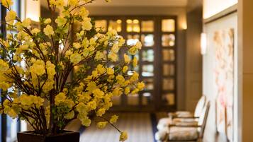 A hallway with a potted plant of yellow flowers, a seating area with chairs, and a door with glass panels.