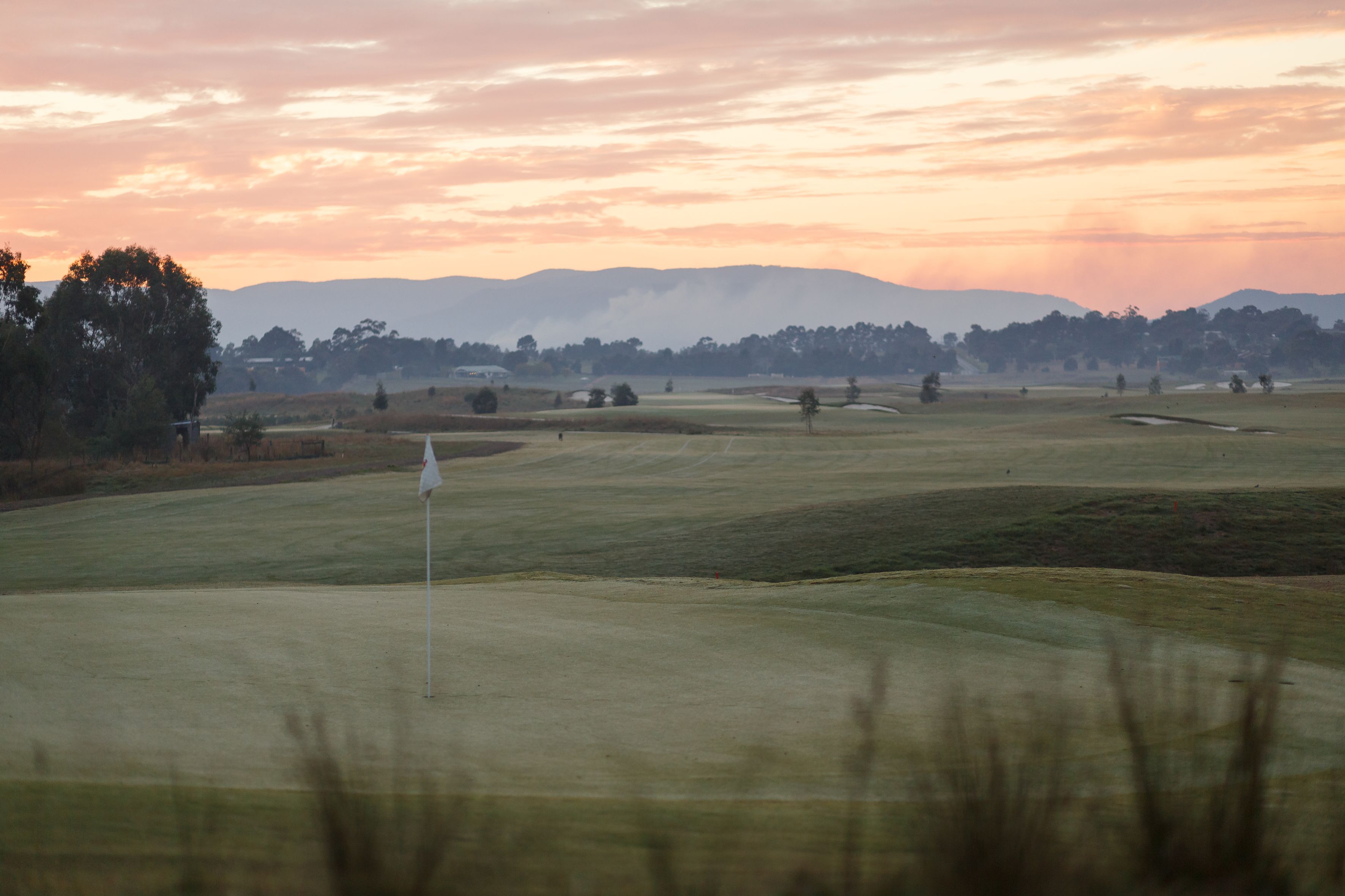 A golf course with a flagstick in the foreground, surrounded by rolling hills and trees.