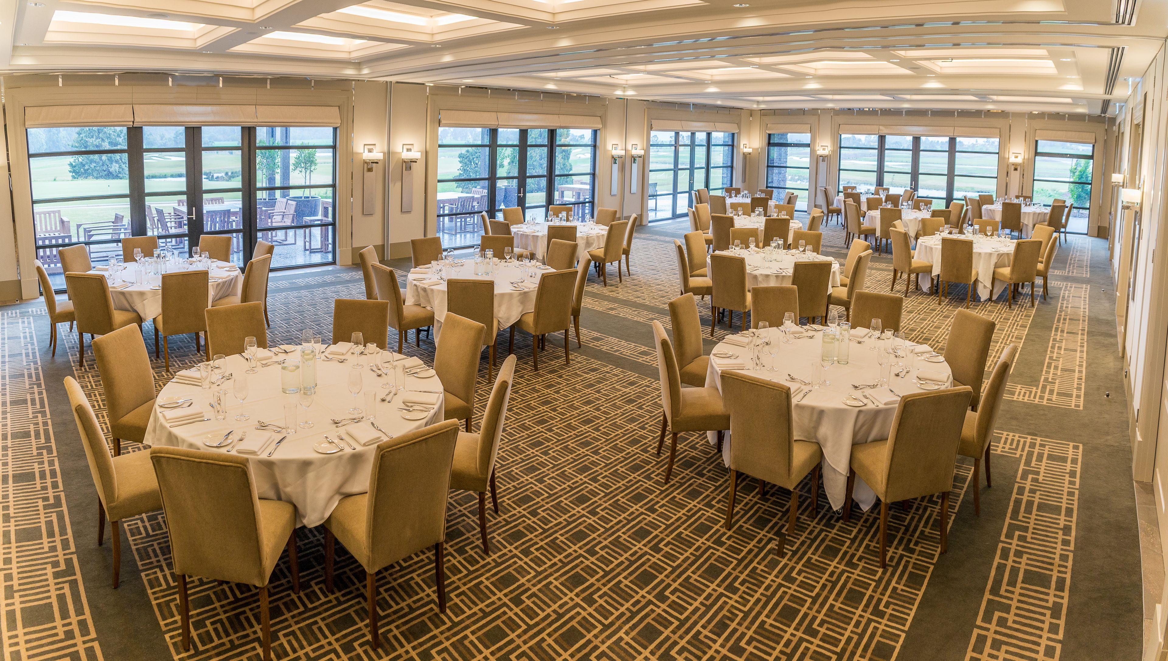A spacious banquet hall with round tables and chairs, large windows offering views of a golf course.