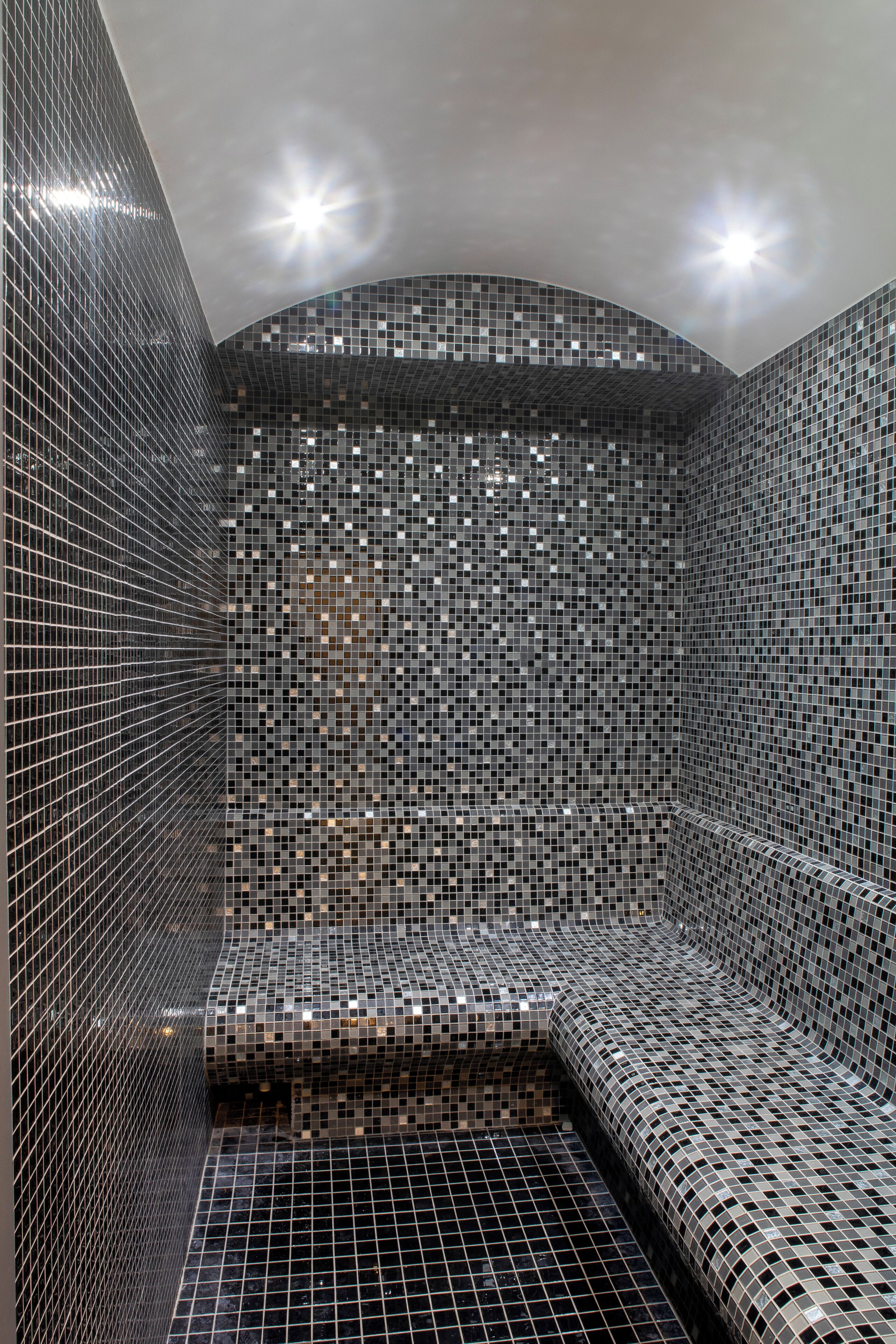 Un ambiente moderno e chiuso, caratterizzato da un muro e un pavimento in mosaico, una panca curva e una struttura in griglia metallica.
