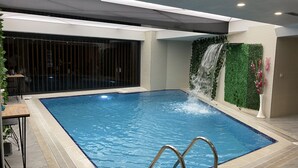 An indoor swimming pool with a waterfall feature, a seating area with a table and chairs, and a decorative wall with plants. - Martinenz Hotel (Istanbul)