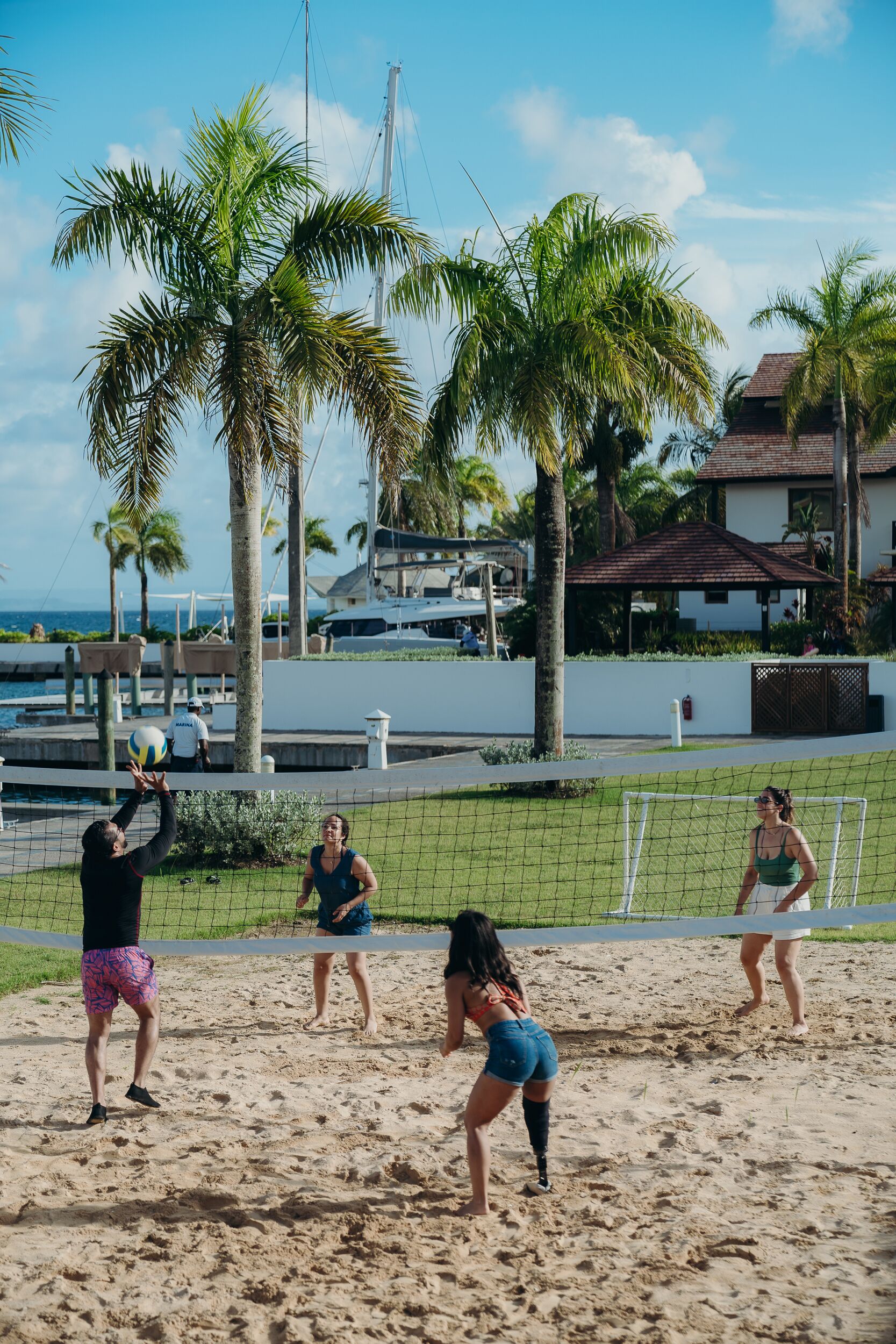 Partido de voleibol de playa con jugadores en la arena, palmeras y un puerto deportivo al fondo.