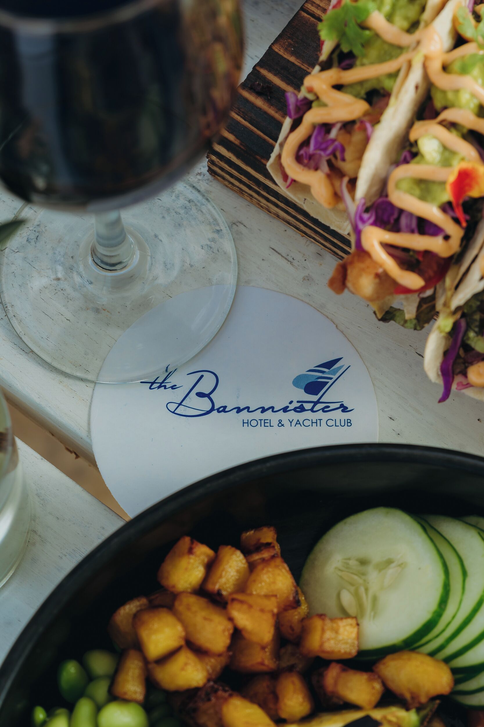 A dining table with a plate labeled "The Bennister Hotel & Yacht Club," a glass of red wine, and a bowl of food including roasted potatoes and sliced cucumbers.