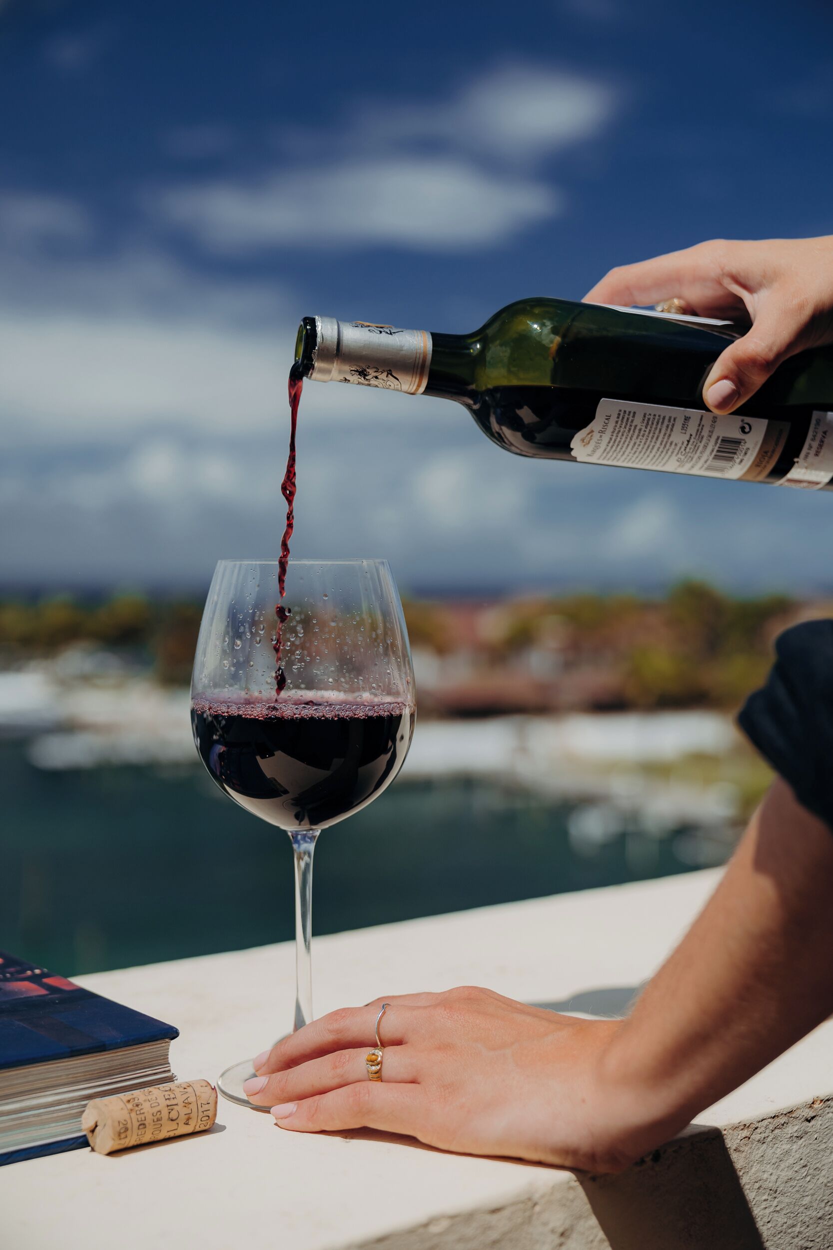A person pouring red wine into a glass, with a book and corkscrew on the table, and a body of water in the background.