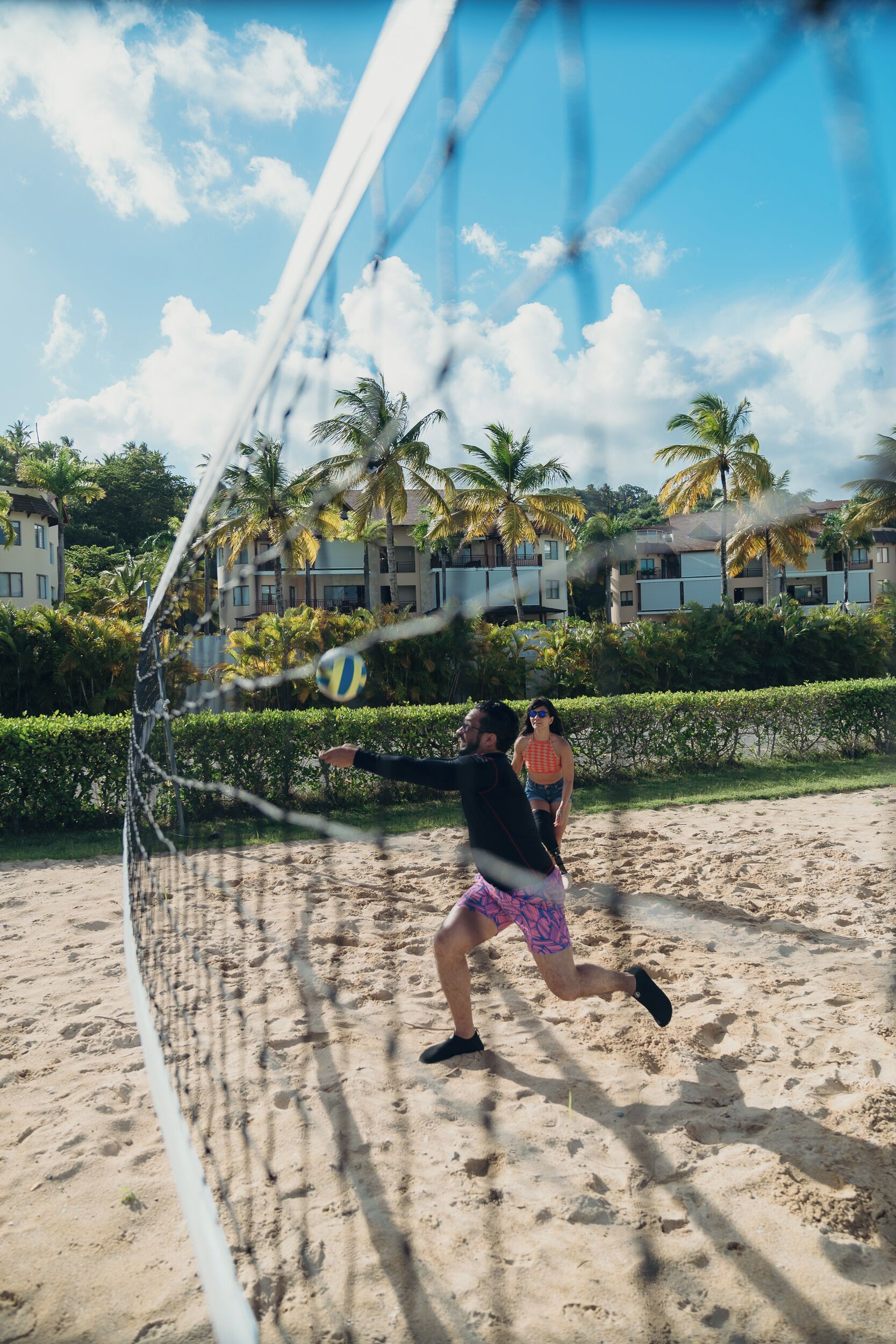 Partido de voleibol de playa con red, palmeras y edificios al fondo.