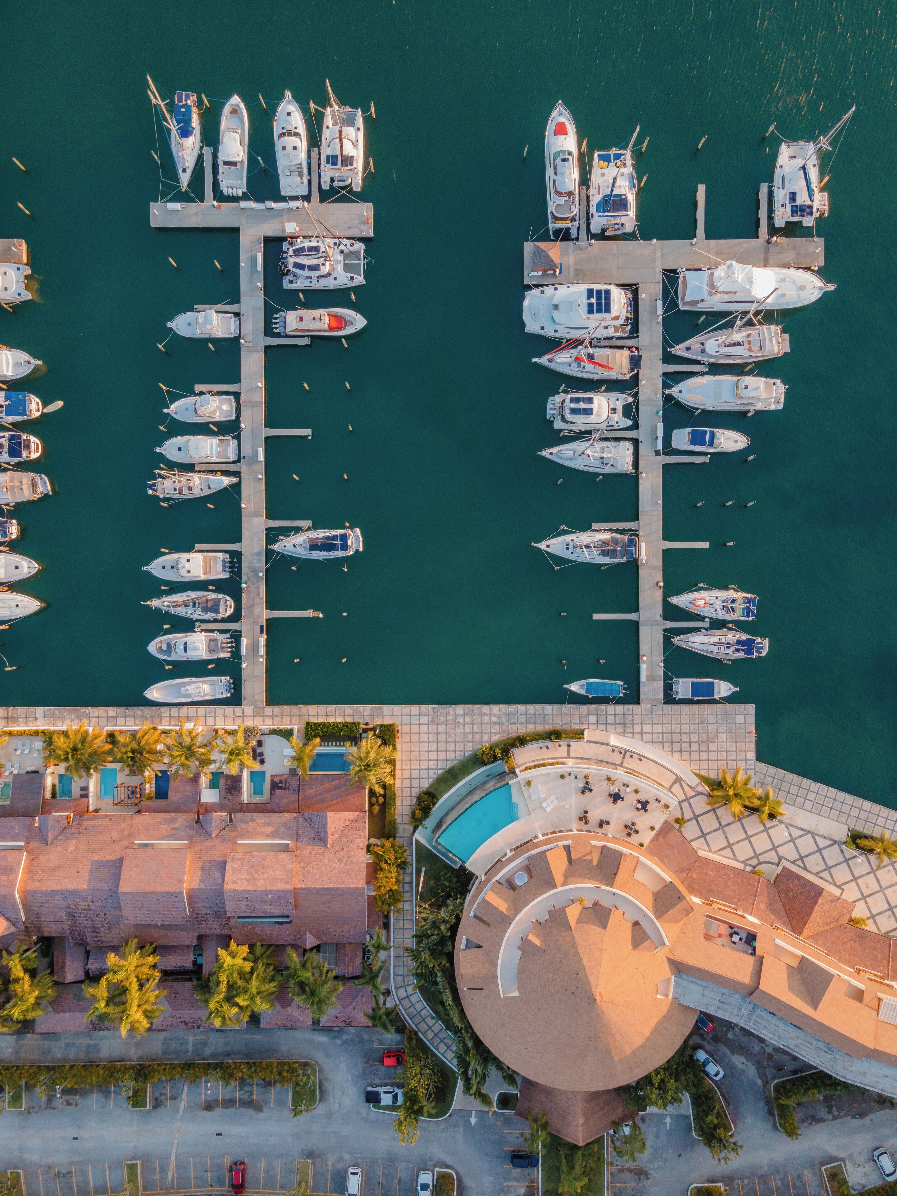 An aerial view of a marina with multiple boats docked at piers, a swimming pool, and a parking area.