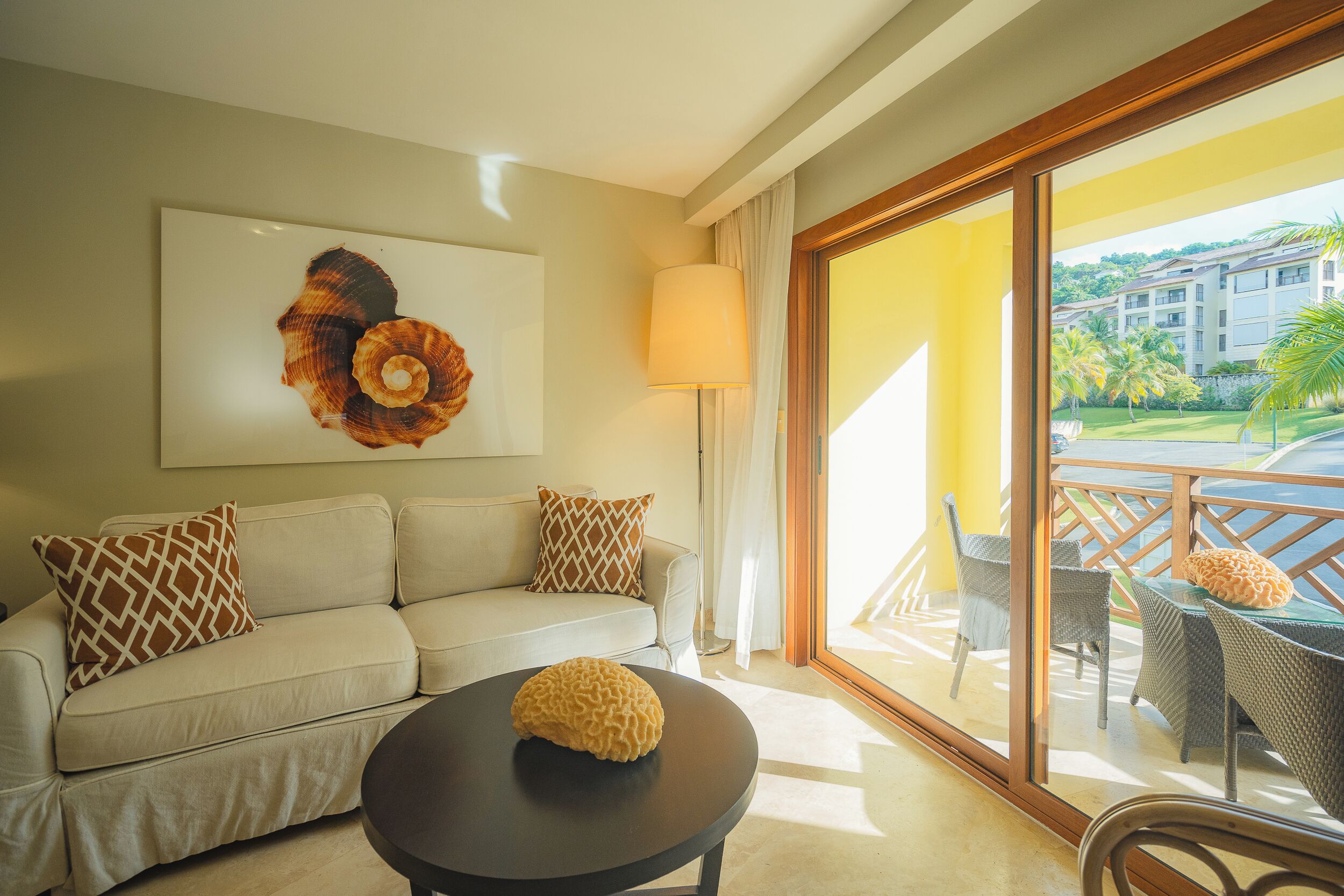 A living room with a sofa, a round table, and a large window with a view of a building and palm trees.
