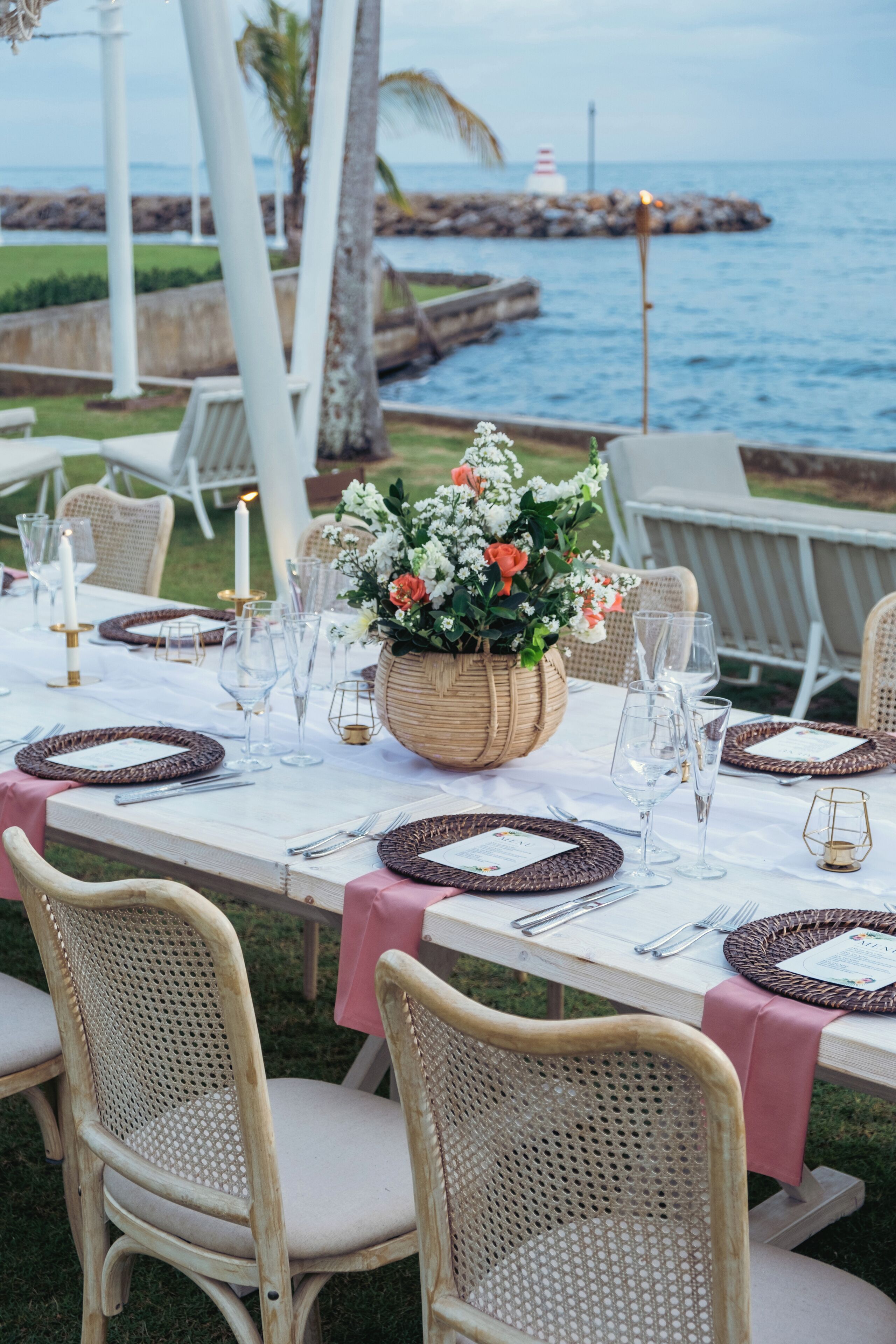 An outdoor dining setup with wicker chairs and tables, a centerpiece with flowers, and a view of the ocean and pier in the background.