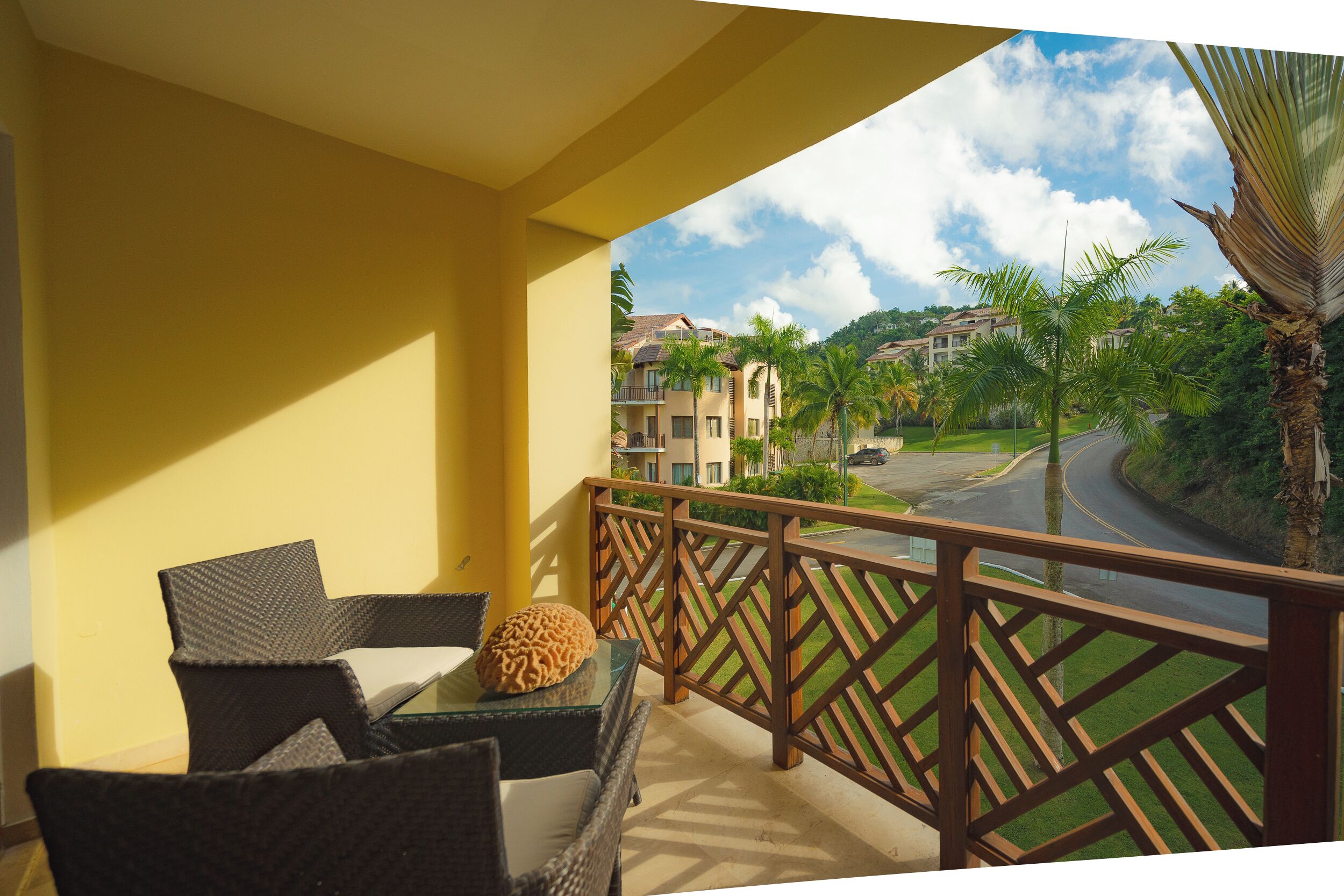 A balcony with wicker furniture and a glass table, overlooking a road and buildings with palm trees.