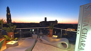 Una terraza en la azotea con mobiliario moderno, plantas en macetas y vistas a la ciudad al atardecer.