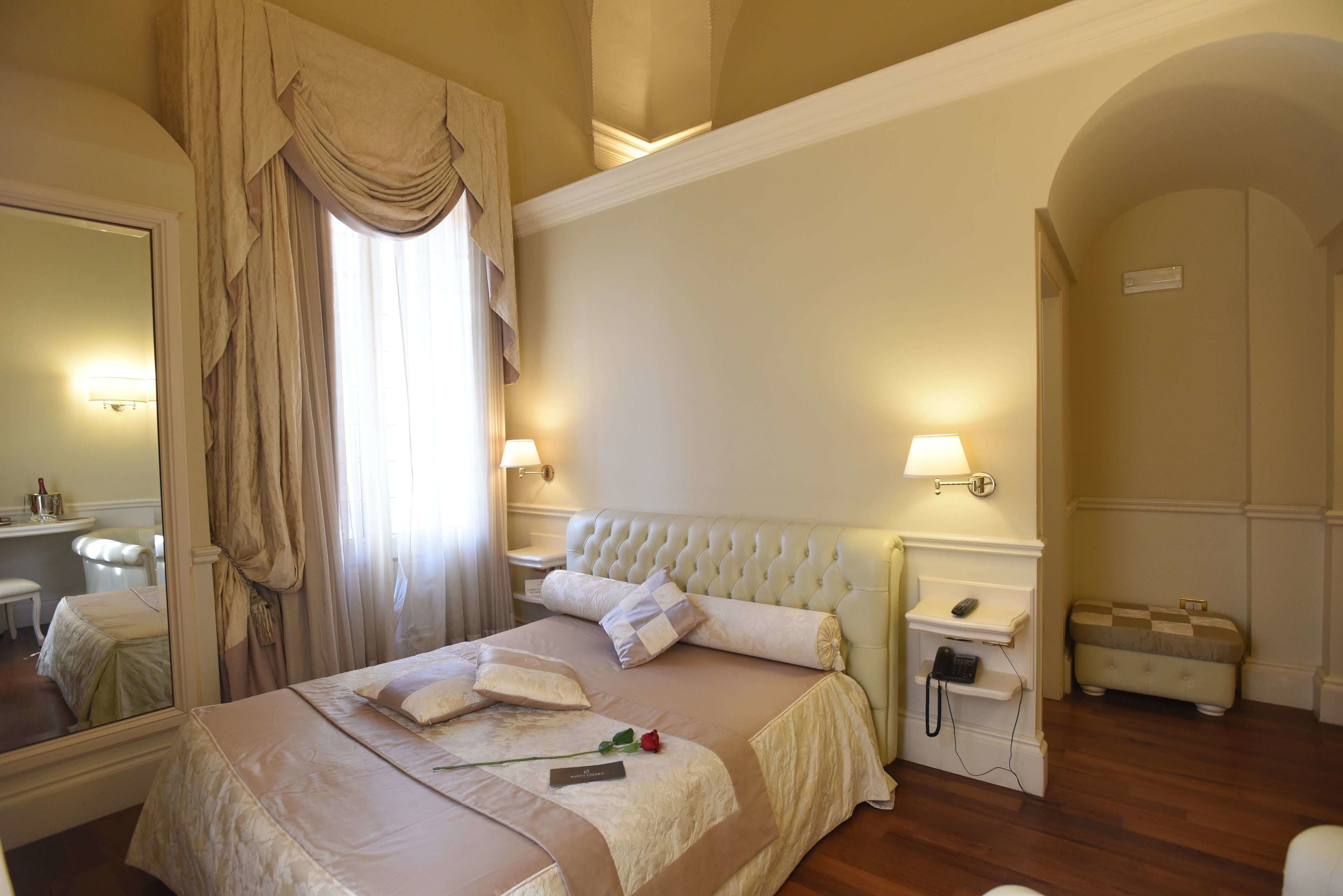 Habitación de hotel con una cama grande, una mesita de noche con lámpara, una mesita más pequeña con teléfono, un espejo y una ventana con cortinas.