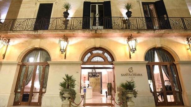 Entrada de hotel con puertas dobles, un letrero que dice "SANTA CHIARA" y un balcón con macetas.