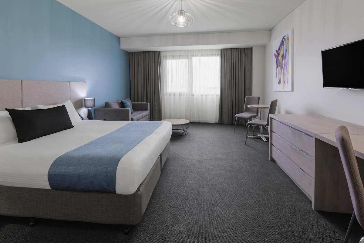 A hotel room with a large bed, a desk, a chair, a television, and a painting on the wall.