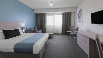 Et hotelværelse med en stor seng, et skrivebord, en stol, et fjernsyn og et billede på væggen.