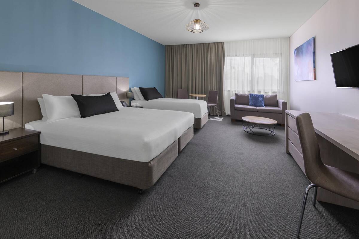 A hotel room with two beds, a desk, a chair, a TV, and a large window with curtains.