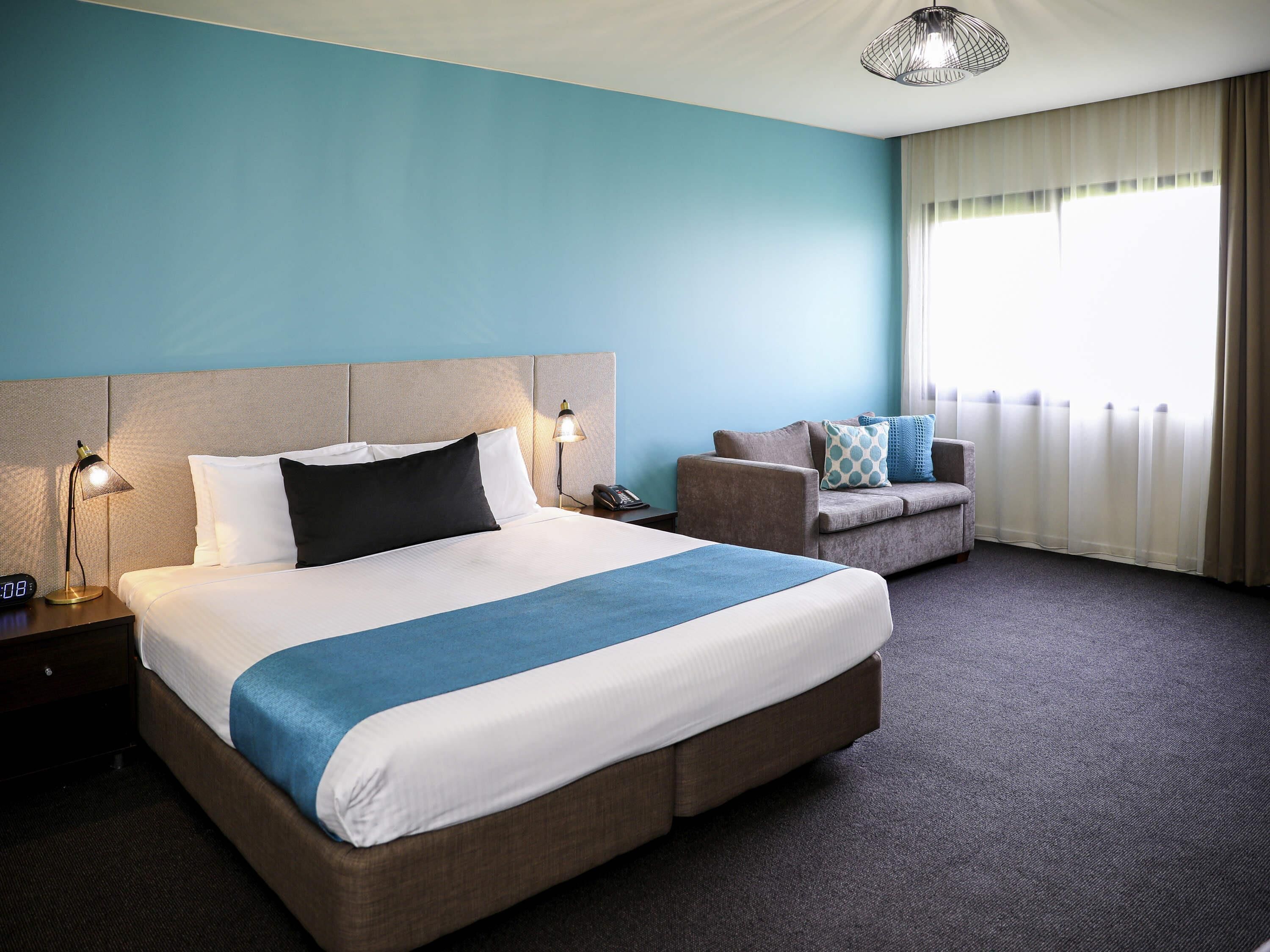 A hotel room with a large bed, a sofa, a nightstand with a lamp, a telephone, and a window with curtains.