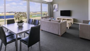A hotel room with a dining area, a sofa set, a flat-screen TV, and large windows with a view of a sports field. - Mantra Melbourne Melton (Cobblebank)