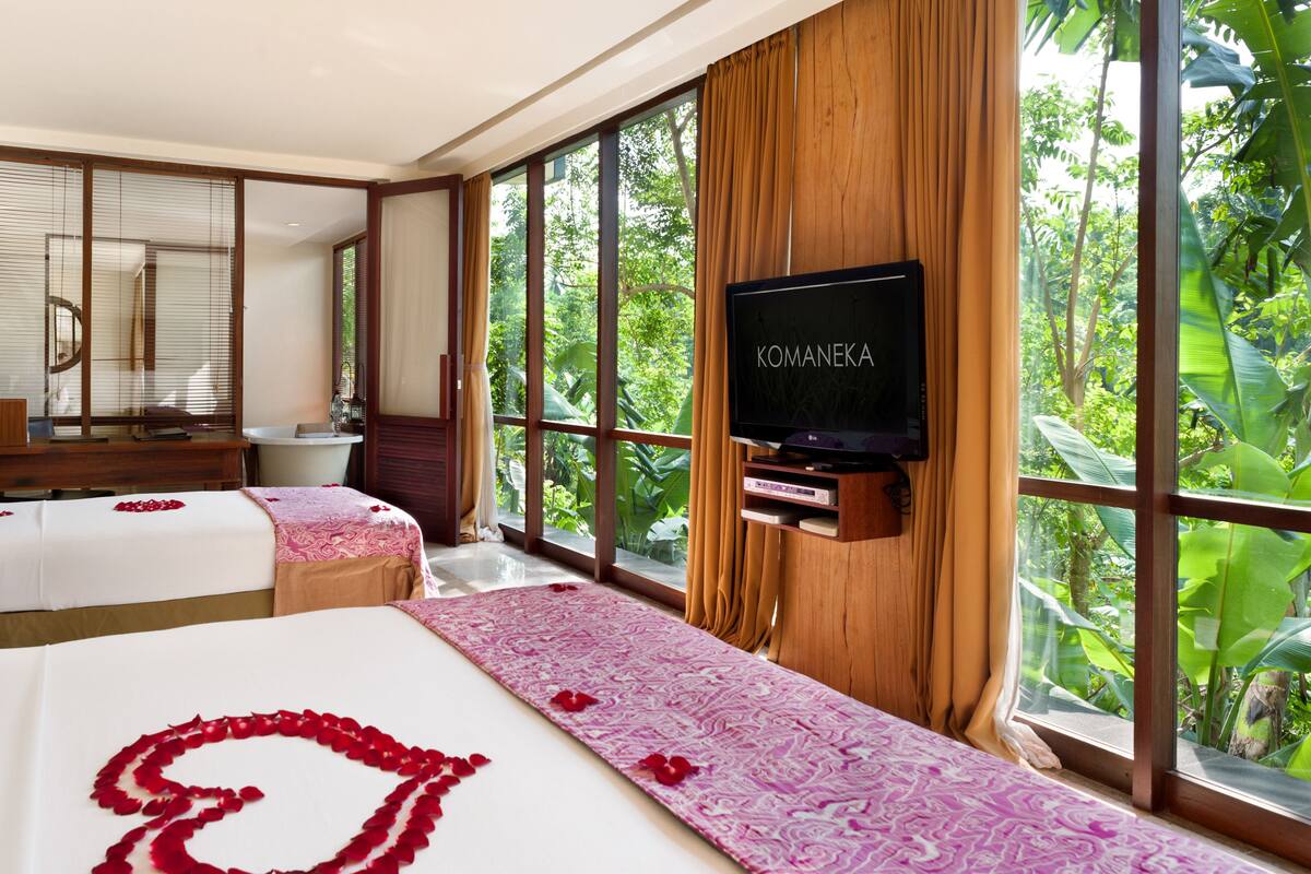 A hotel room with two beds, a TV, and large windows with a view of greenery.