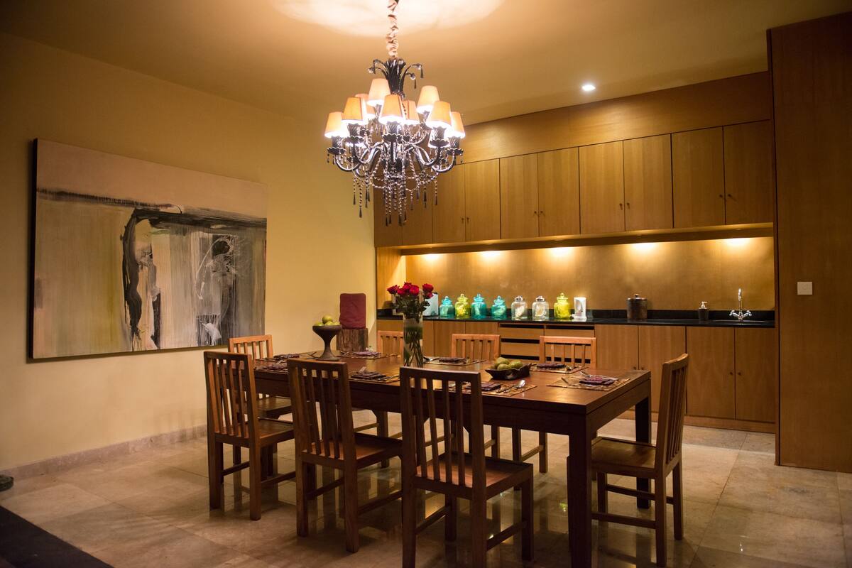 A dining area with a wooden table and chairs, a chandelier, and a kitchen in the background.