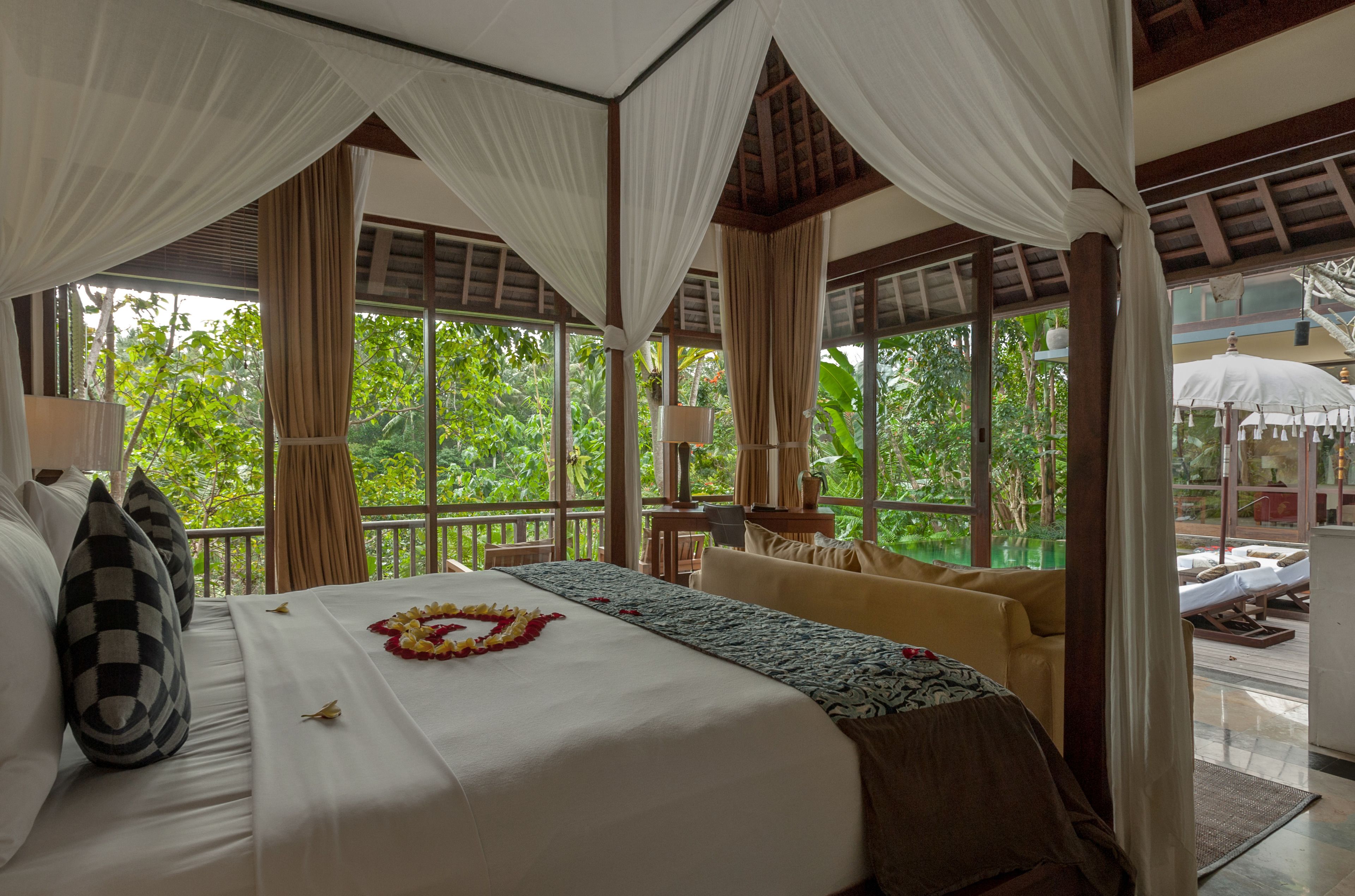 A spacious room with a large bed, a sofa, and a small table. There are curtains and a view of greenery outside.