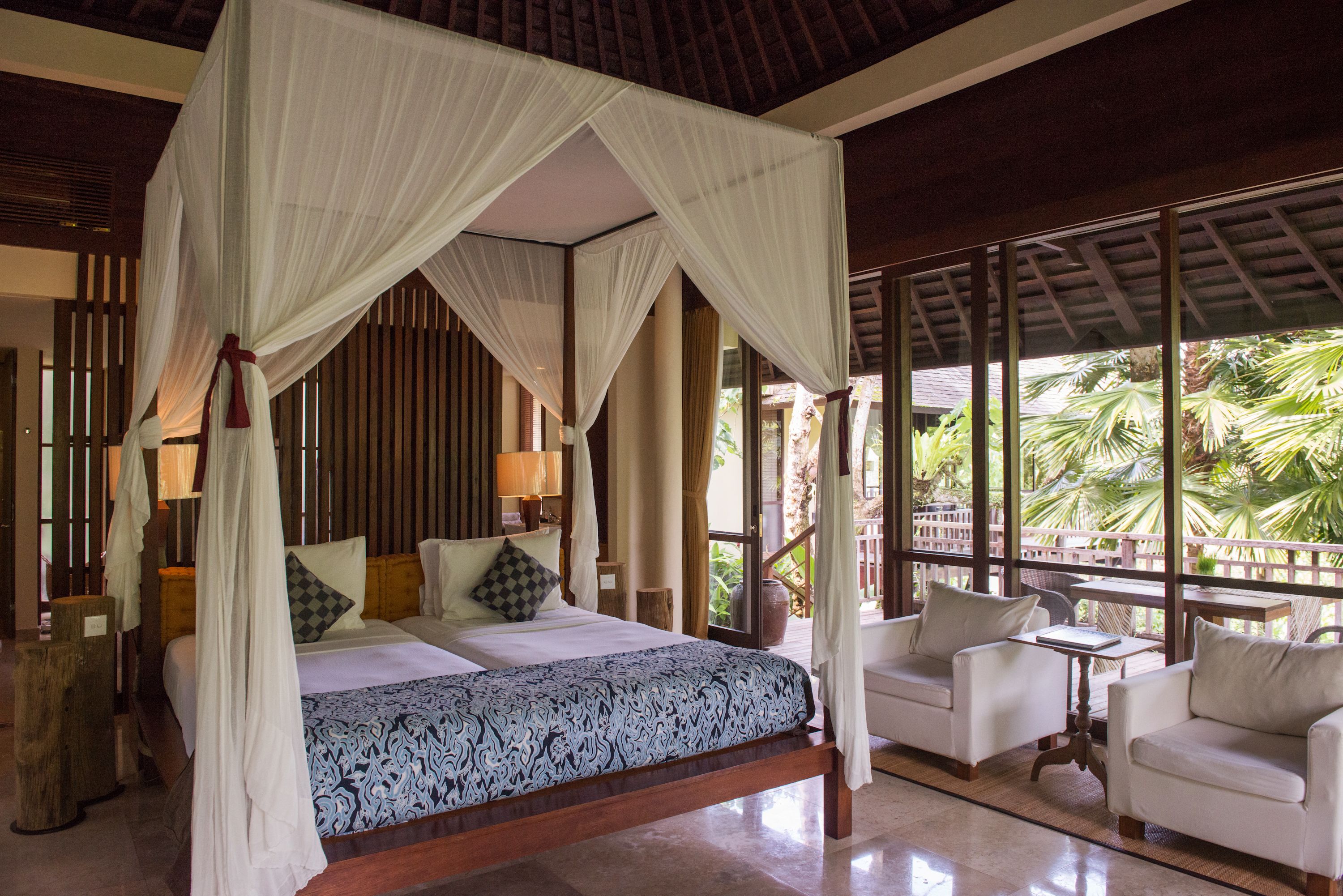 A four-poster canopy bed with a patterned bedspread, two bedside tables with lamps, and a view of palm trees through large windows.