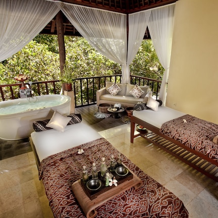 A spa suite with a massage table, a massage table with a patterned bedspread, a bathtub, and a small table with bottles and a bowl.