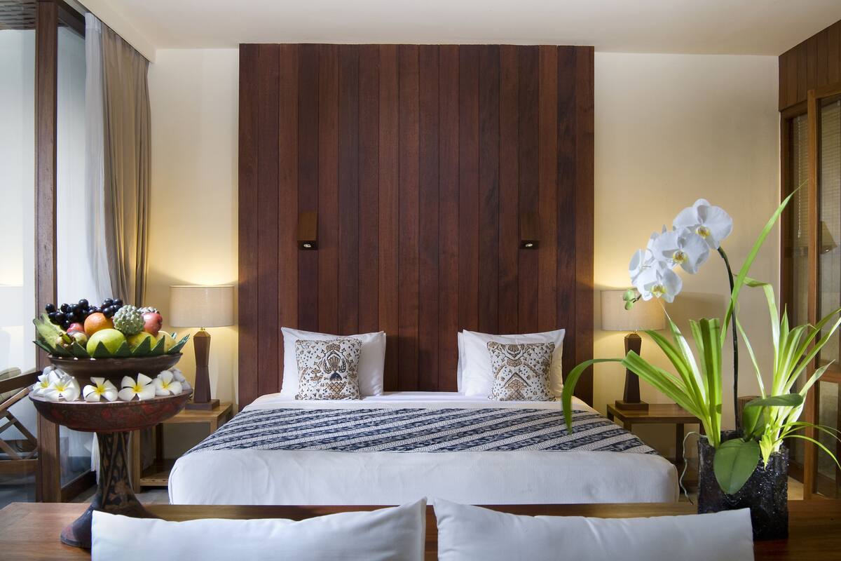 A hotel room with a large bed, two pillows, a fruit bowl, a potted orchid, and a nightstand with a lamp.