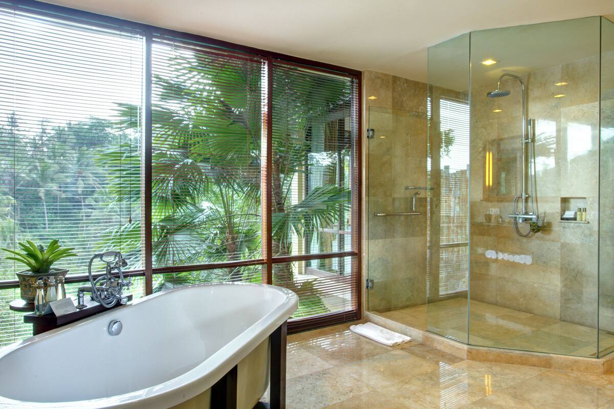 A modern bathroom with a glass-enclosed shower, a freestanding bathtub, and a large window with blinds.