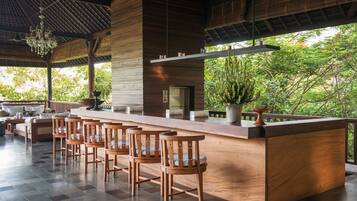 A modern bar area with wooden chairs and a long counter, surrounded by large windows with a view of greenery.
