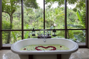 A bathtub with a heart-shaped flower arrangement, set against a backdrop of lush greenery visible through large windows.