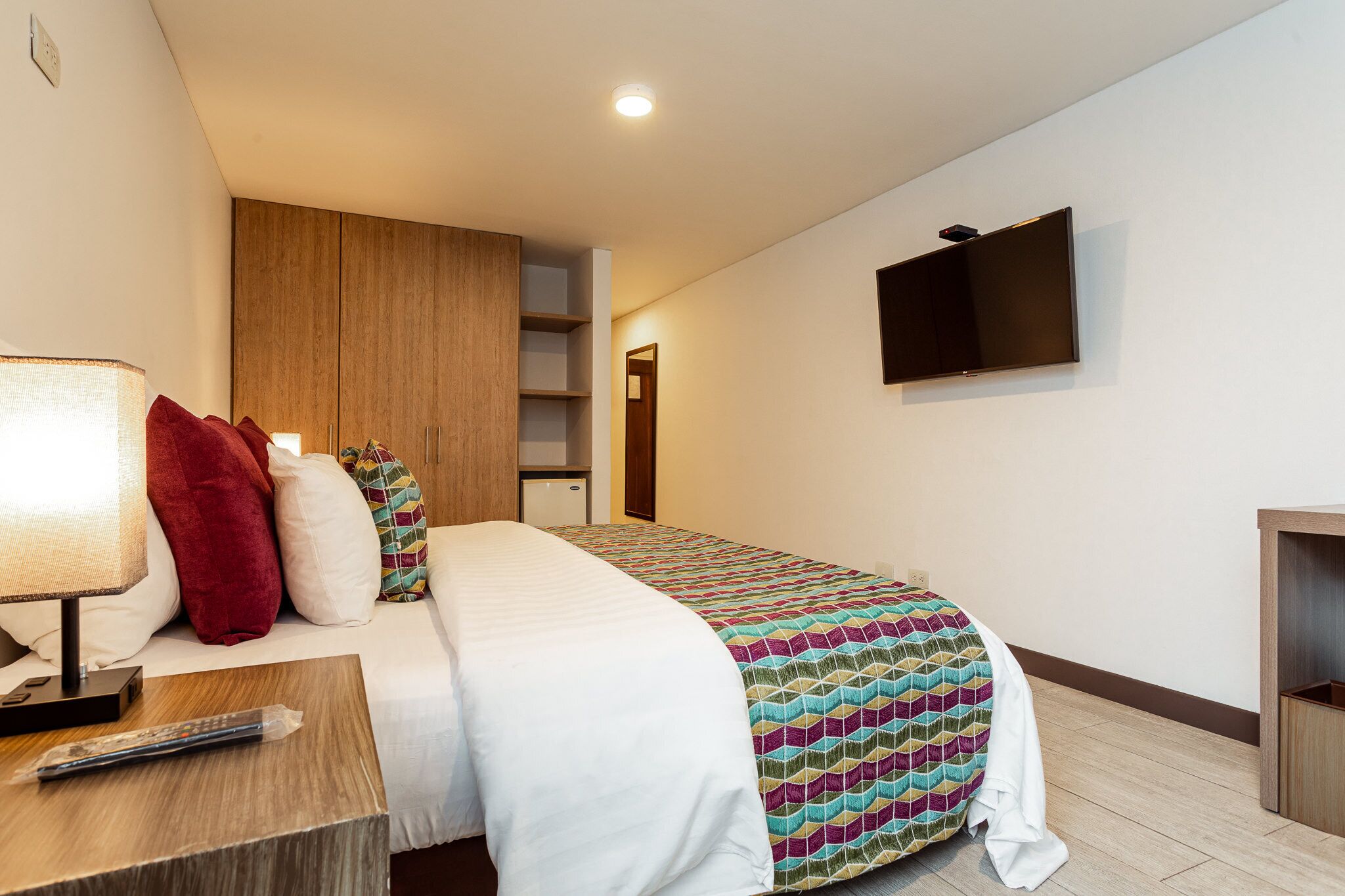 A hotel room with a bed, a TV mounted on the wall, a bedside table with a lamp, and a wooden wardrobe.