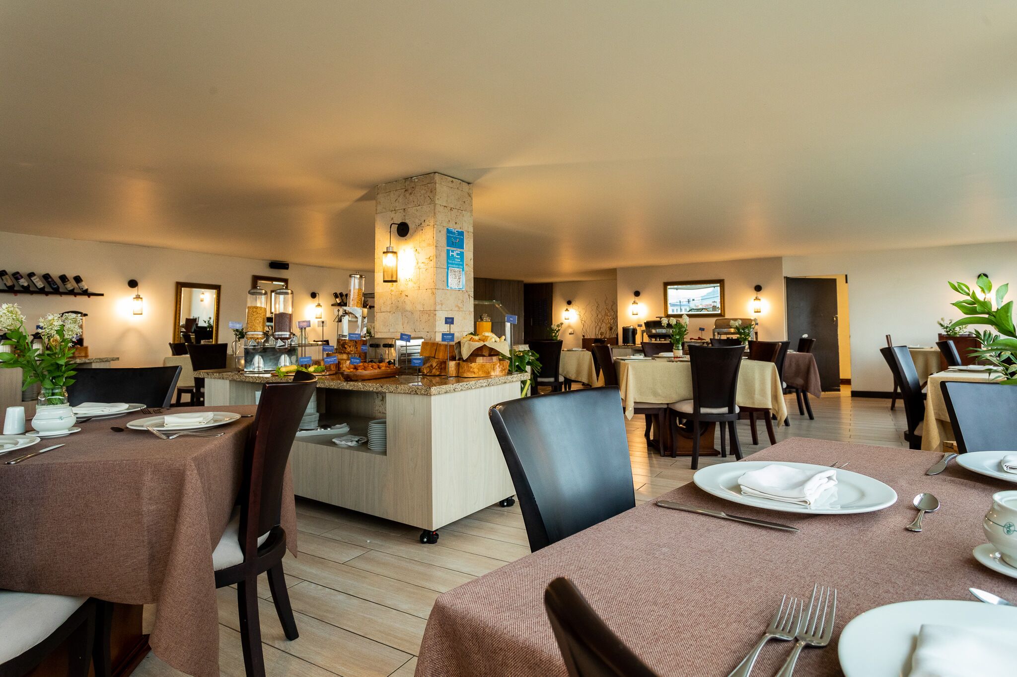 A restaurant with multiple tables set for dining, a central bar area, and various plants and decorations.