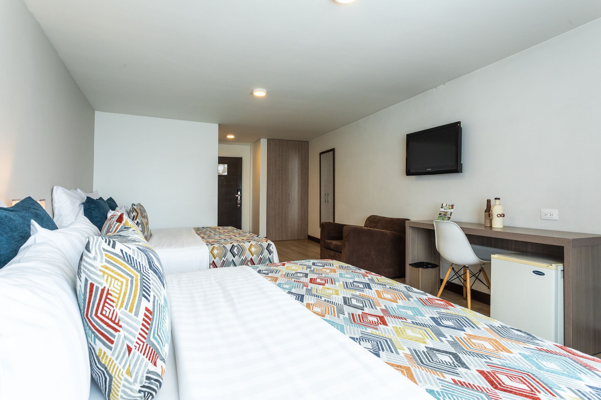 A hotel room with two beds, a desk, a chair, a TV, and a small fridge.