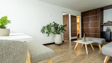 A modern living room with a white table, grey sofas, wooden flooring, and large potted plants.