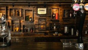 A bar area with wooden shelves displaying various bottles of alcohol, a sign for BBC Cerveza Fresca de Barril, and a coffee maker. - Hotel Carretero (Manizales)