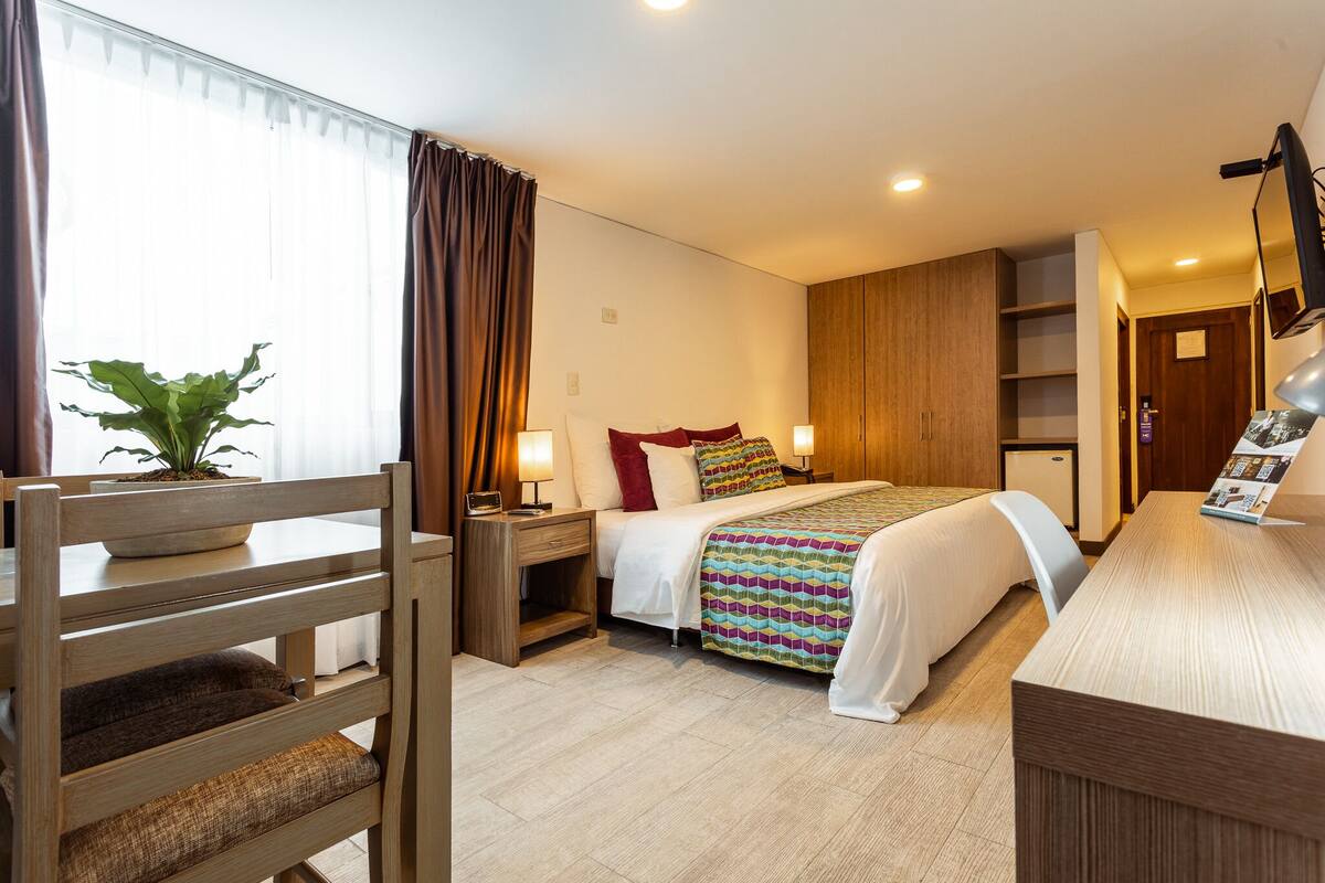 A hotel room with a bed, bedside tables, a desk, a chair, a potted plant, and a television.