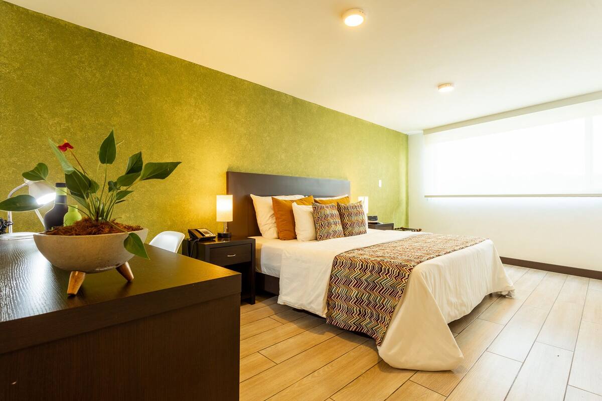A hotel room with a bed, bedside table, lamp, and a potted plant.