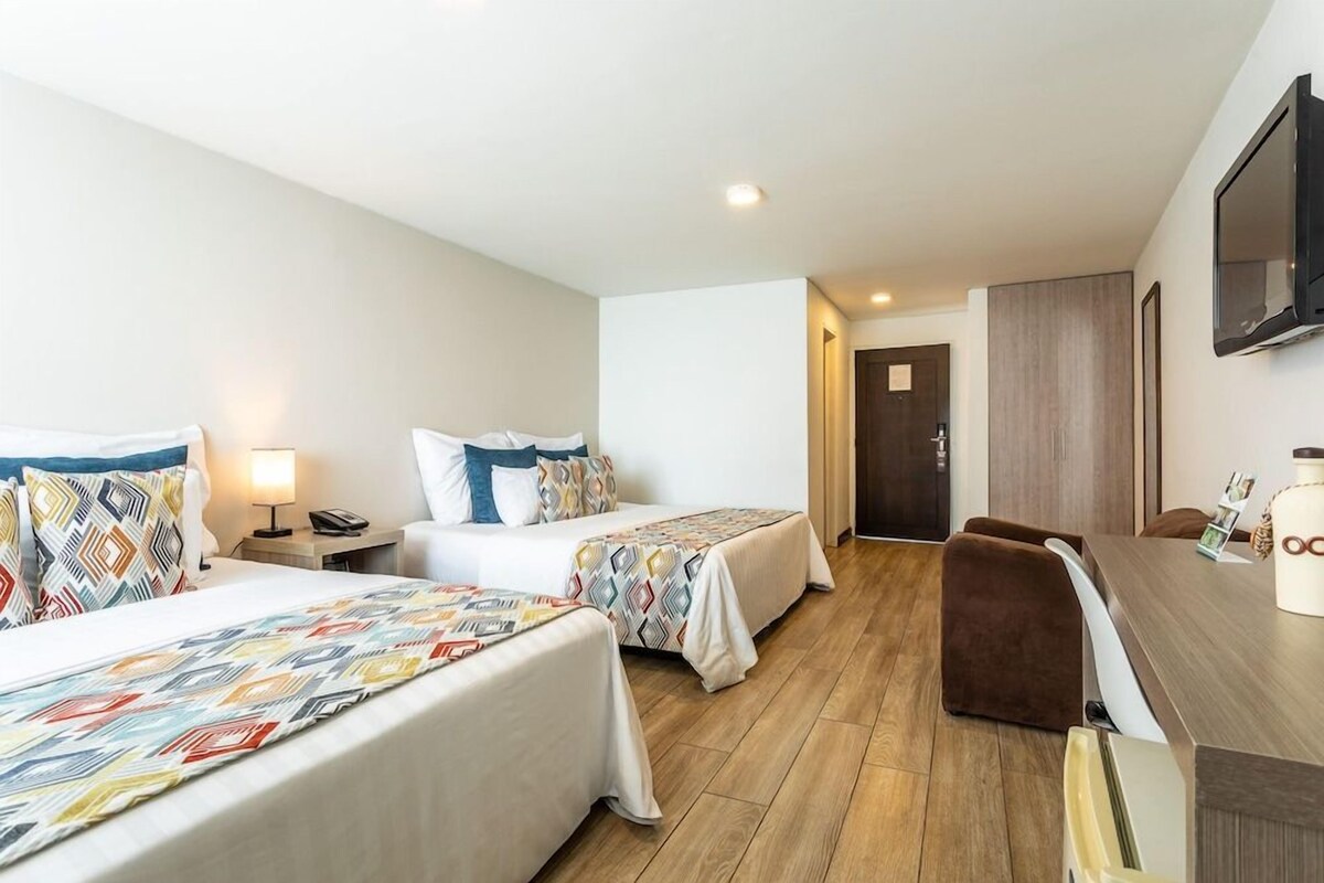 A hotel room with two beds, a desk, a chair, a TV, and a door leading to another room.