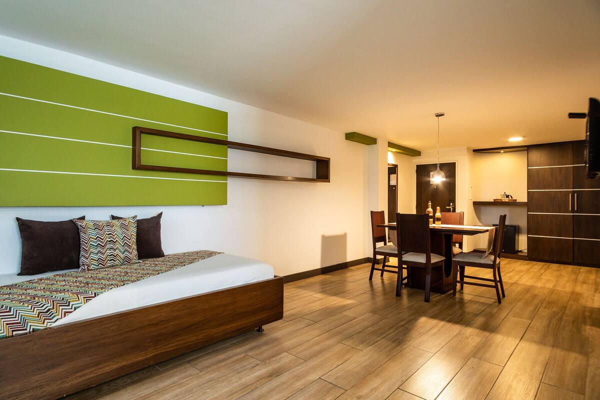 A modern hotel room with a bed, a dining area, and a kitchen.