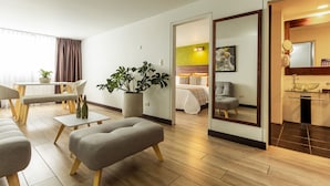 A modern hotel room with a wooden floor, a dining area with a table and chairs, a seating area with a sofa and ottoman, a large mirror, and a bathroom with a glass sink. - Hotel Carretero (Manizales)
