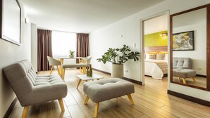 A modern hotel room with a sofa, ottoman, dining table, chairs, a potted plant, a bed, and a mirror reflecting the room. - Hotel Carretero (Manizales)