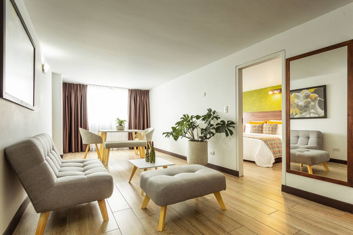 A modern hotel room with a sofa, ottoman, dining table, chairs, a potted plant, a bed, and a mirror reflecting the room.