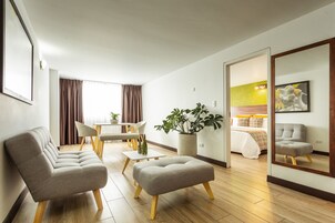 A modern hotel room with a sofa, ottoman, dining table, chairs, a potted plant, a bed, and a mirror reflecting the room.