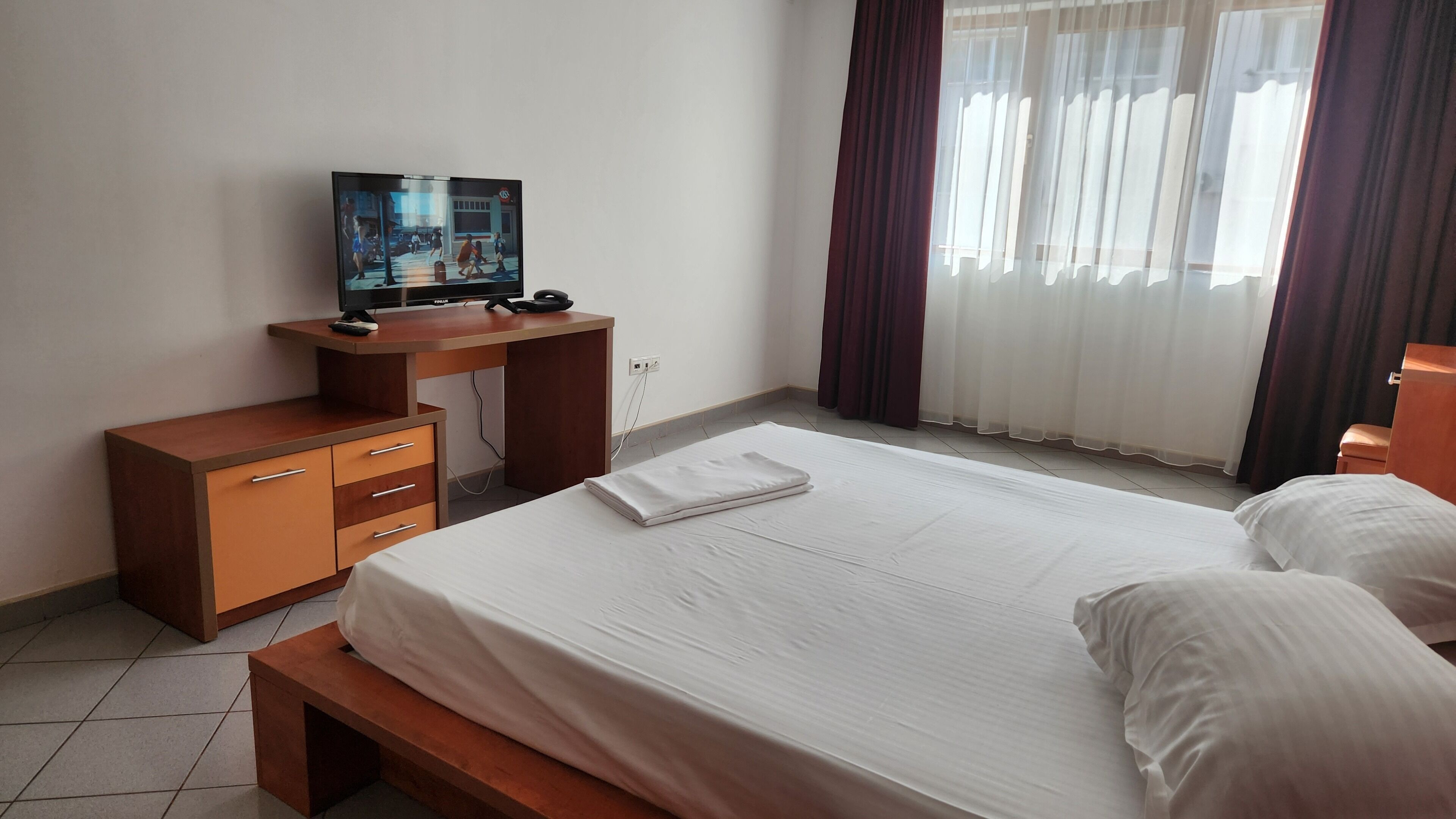 A hotel room with a bed, a desk with a television, a bench, and a window with curtains.