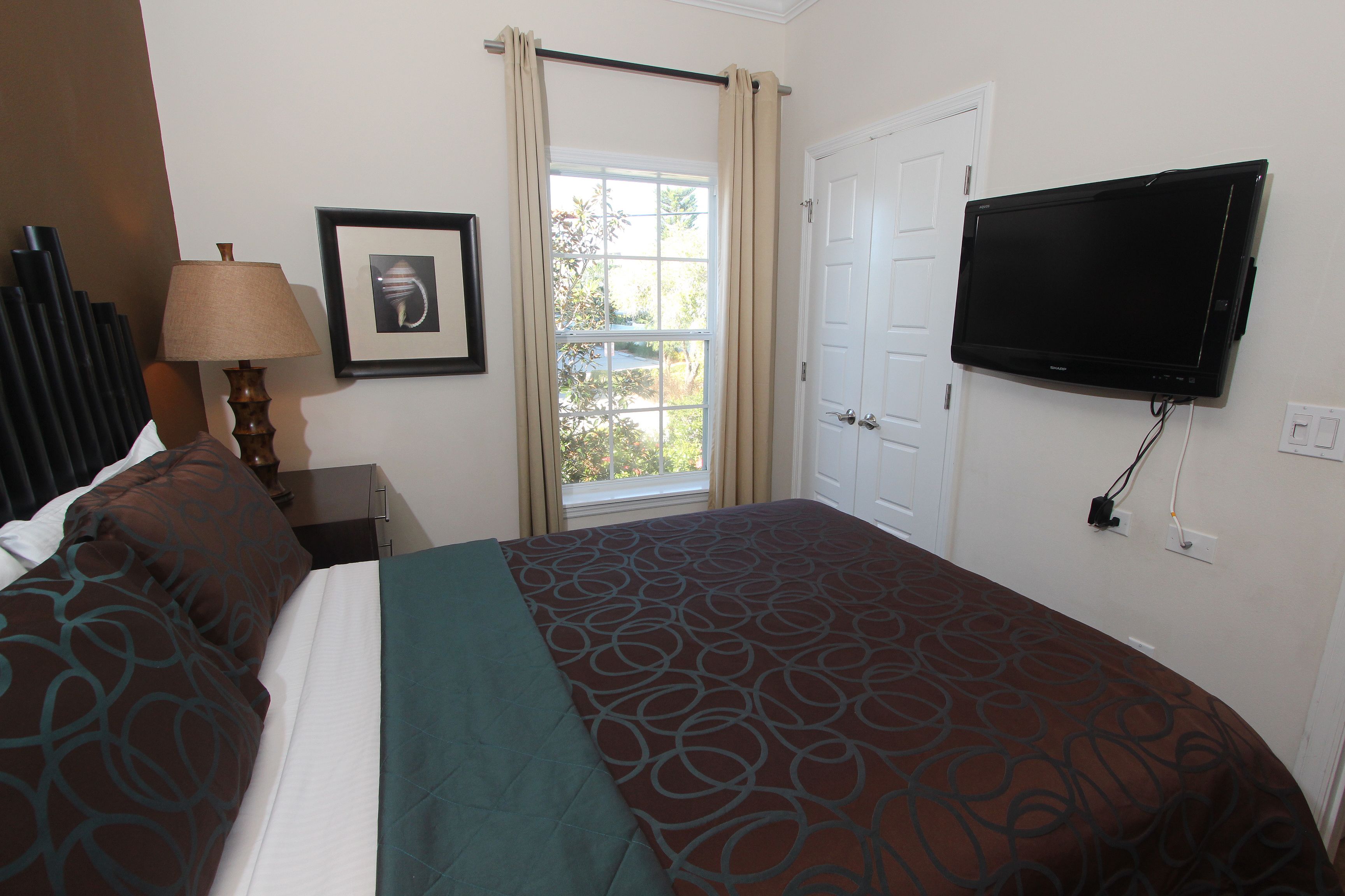 A hotel room with a bed, a TV mounted on the wall, a window with curtains, and a framed picture on the wall.